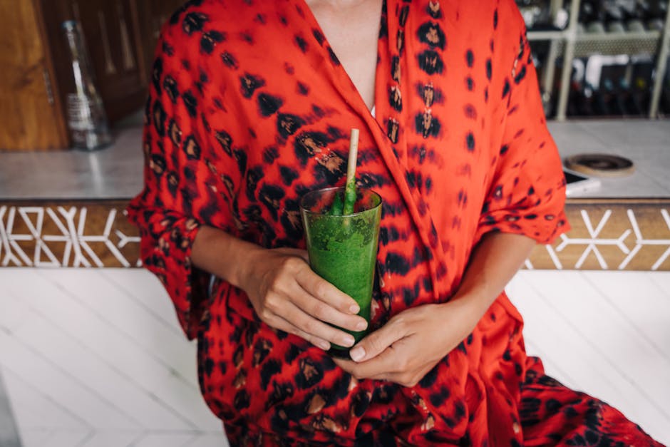 Close-up of a person holding a green smoothie in a chic bar setting, wearing a vibrant red robe.