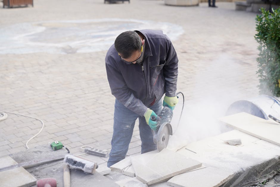 A construction worker uses a power saw to cut stone tiles outdoors, creating dust as he works.