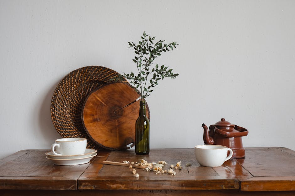 A rustic still life showcasing a teapot, cups, and a bottle with branches on a wooden table.