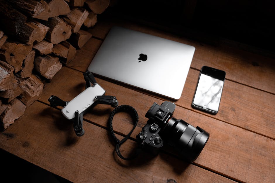 A flat lay of modern technology devices including a laptop, camera, drone, and smartphone on a wooden table.